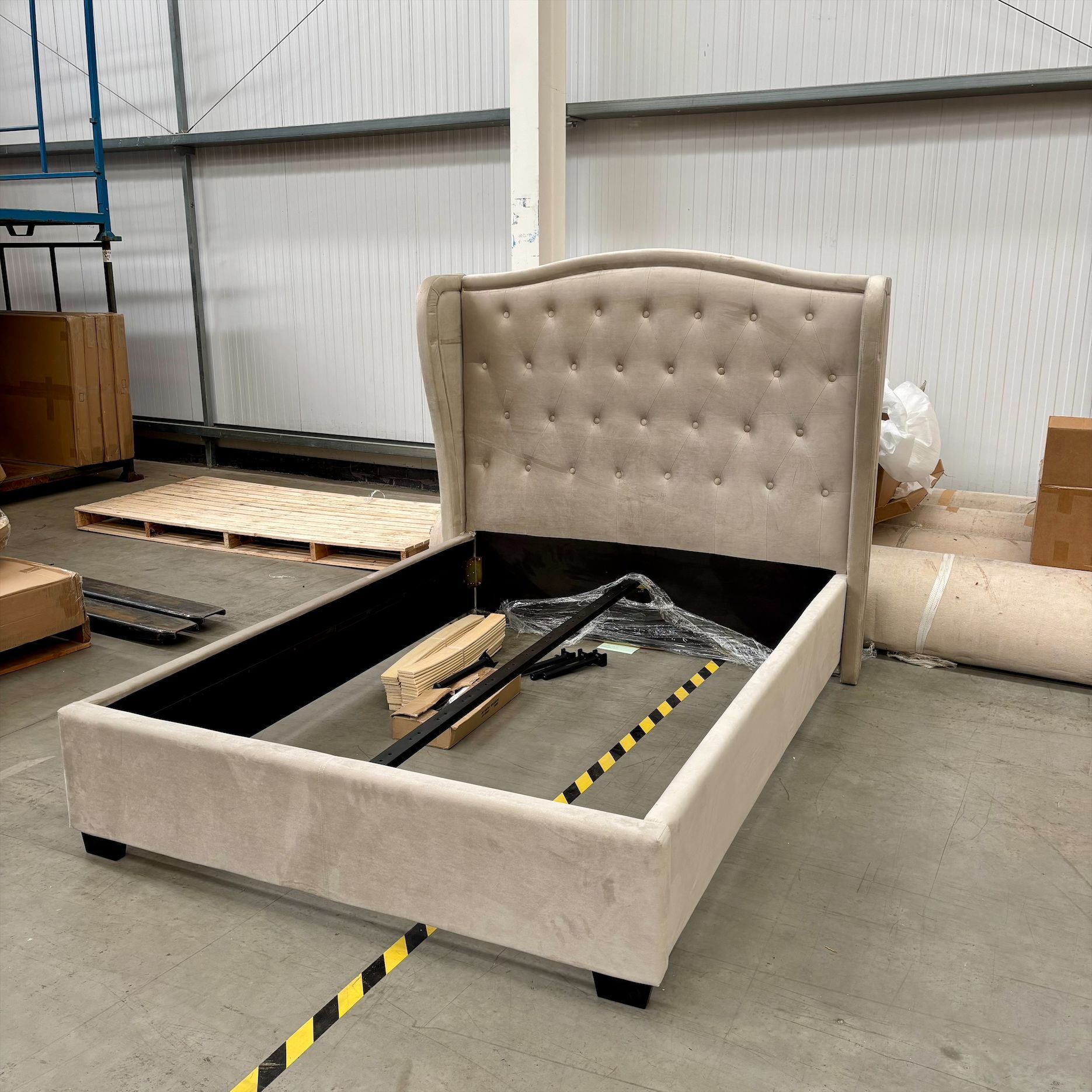 Beige upholstered bed frame in a warehouse, with tools inside the open base.
