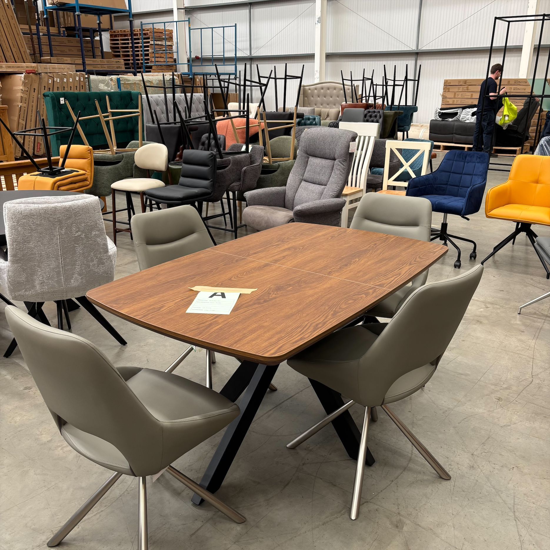 Furniture showroom with a wooden table, mixed chairs, and a gray rug in a warehouse-style space