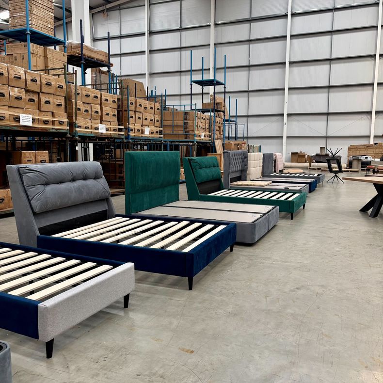 Warehouse aisle with several upholstered bed frames and mattresses in blue, gray, and green.