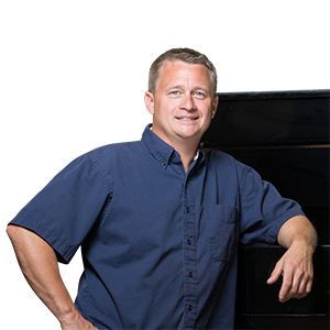 Man in a blue shirt leaning against a black cabinet, smiling.