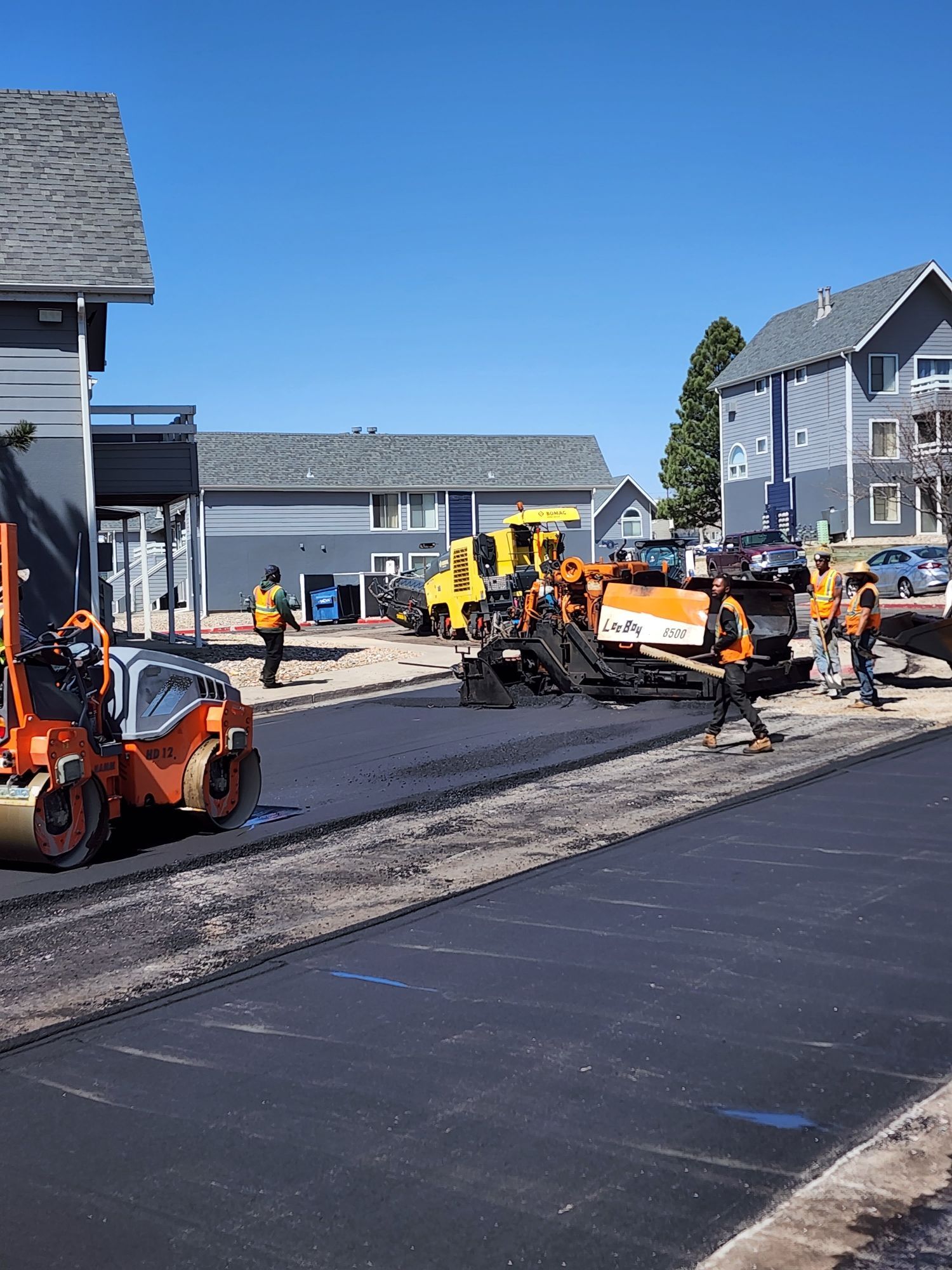 Asphalt paving construction in progress; workers and machinery on new road surface near buildings.