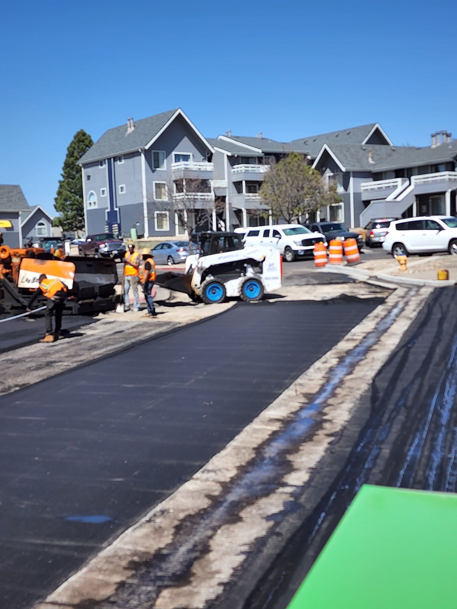 Paving a road in front of apartment buildings. Workers in safety vests operate machinery, clear blue sky.