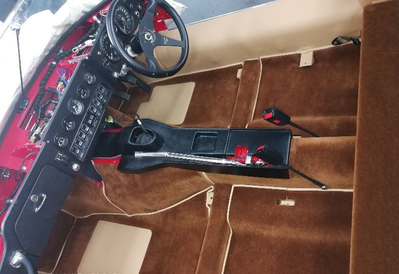 Interior of a classic car with brown carpet and beige trim. Black dashboard and steering wheel visible.