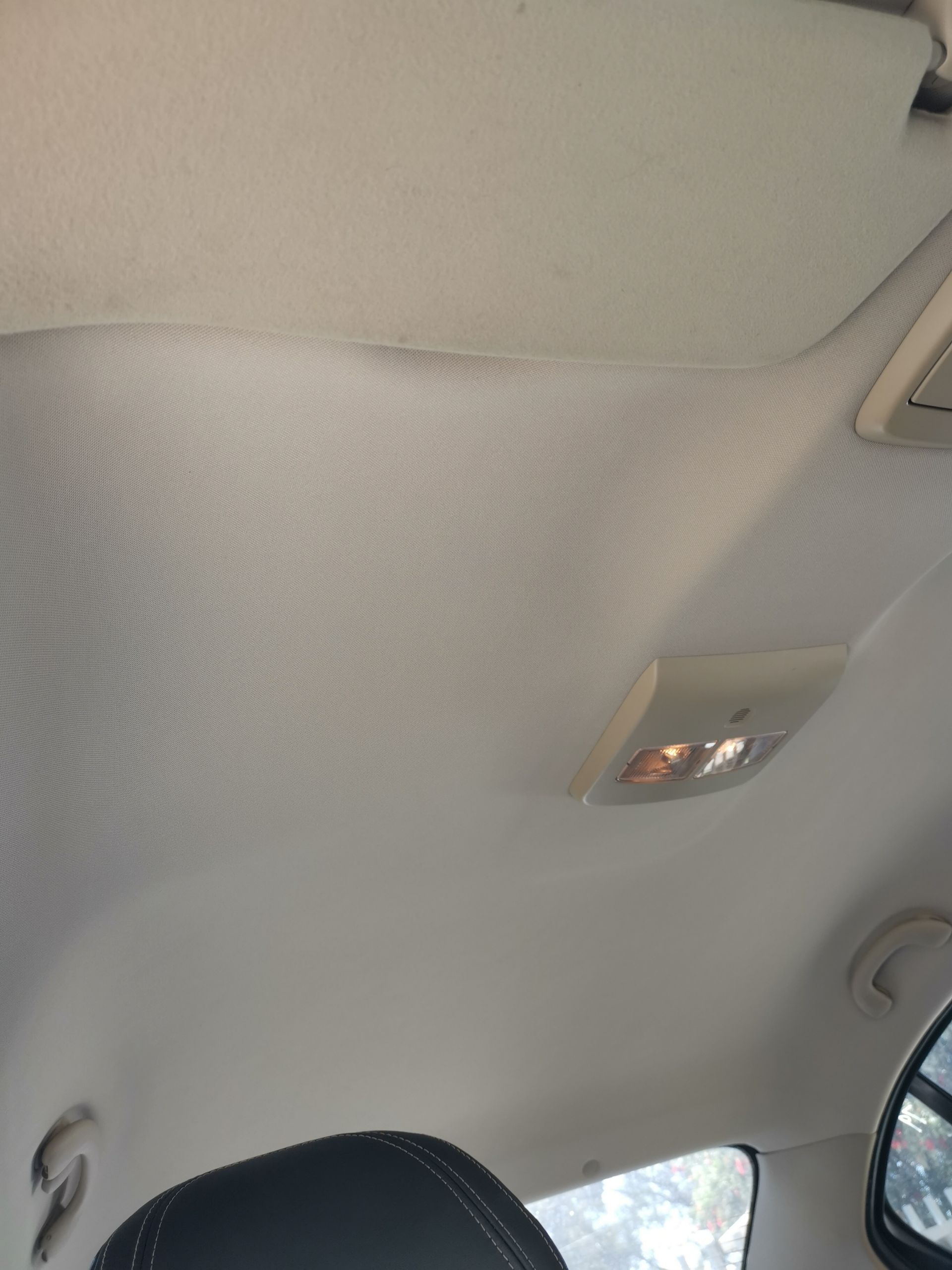 Car's interior ceiling, light gray fabric with stains, overhead light and door handle visible.