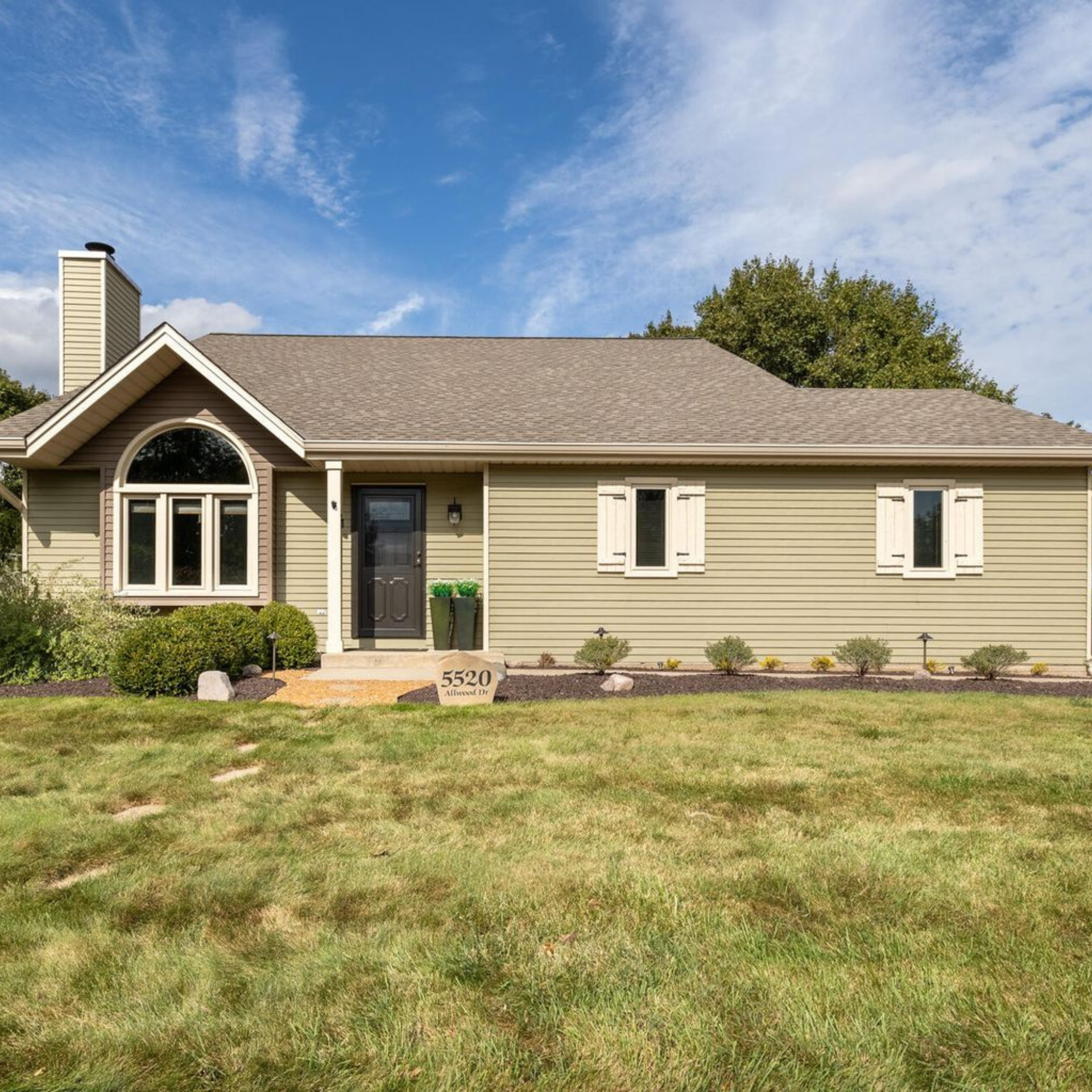 Green ranch house with a gray roof, black door, and white window trim under a blue sky.