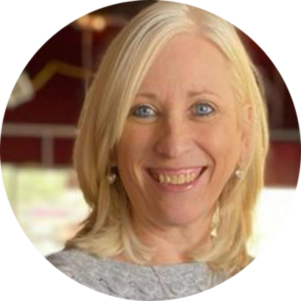 Blonde woman with blue eyes smiles broadly; outdoors, wearing gray shirt and heart earrings.