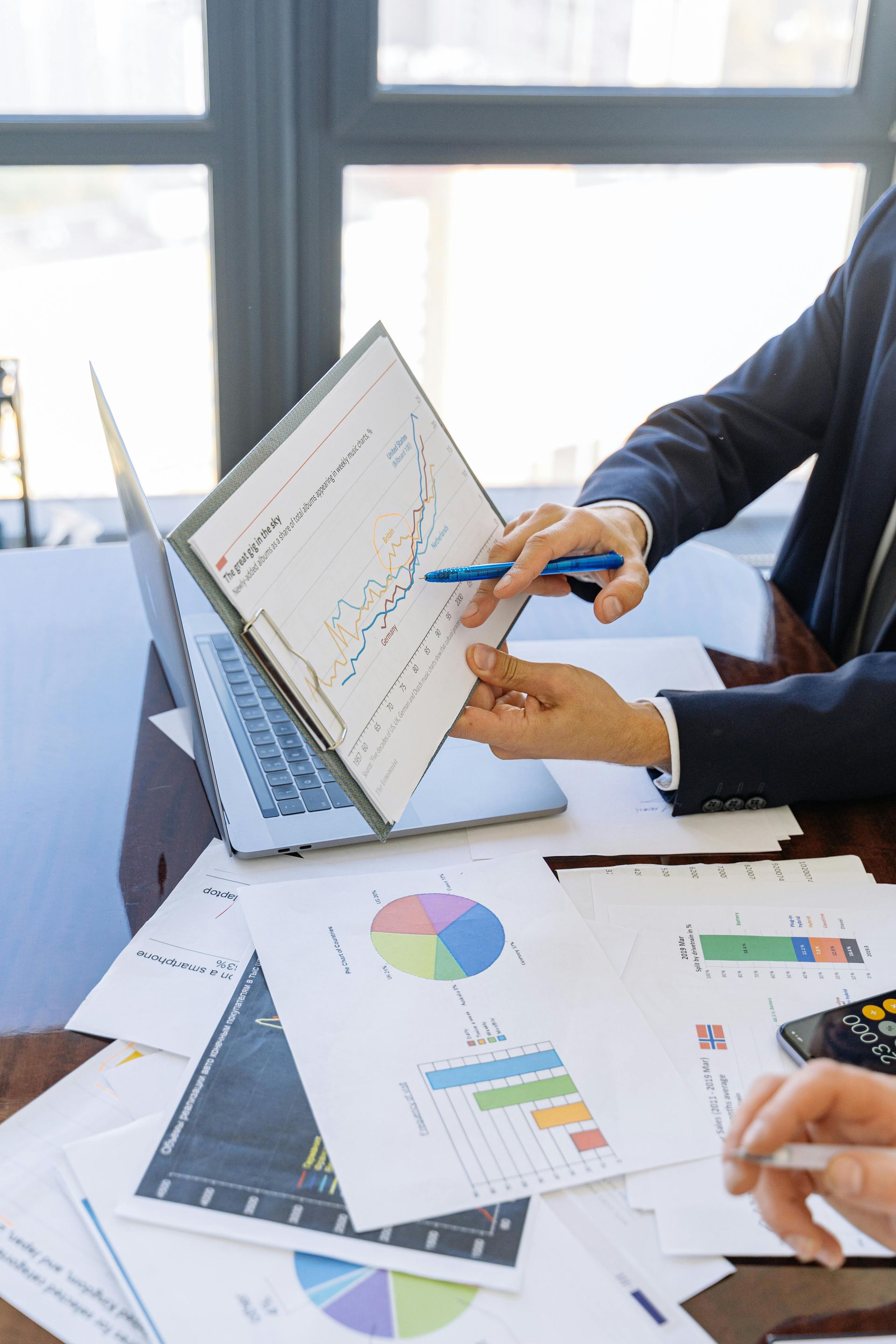 Person in suit points to financial chart with pen; graphs, laptop, and papers on table.