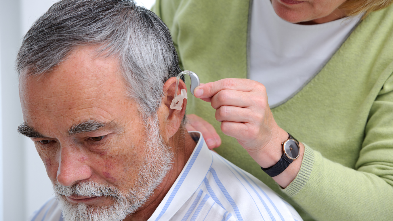 Woman adjusting hearing aid on an older man's ear. Indoors, light skin tones, neutral background.