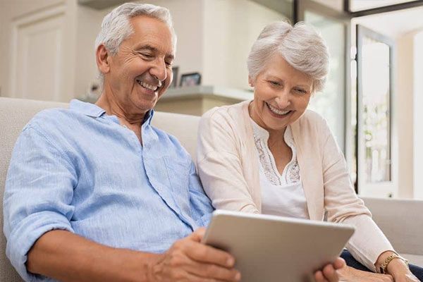 An elderly couple is sitting on a couch looking at a tablet.