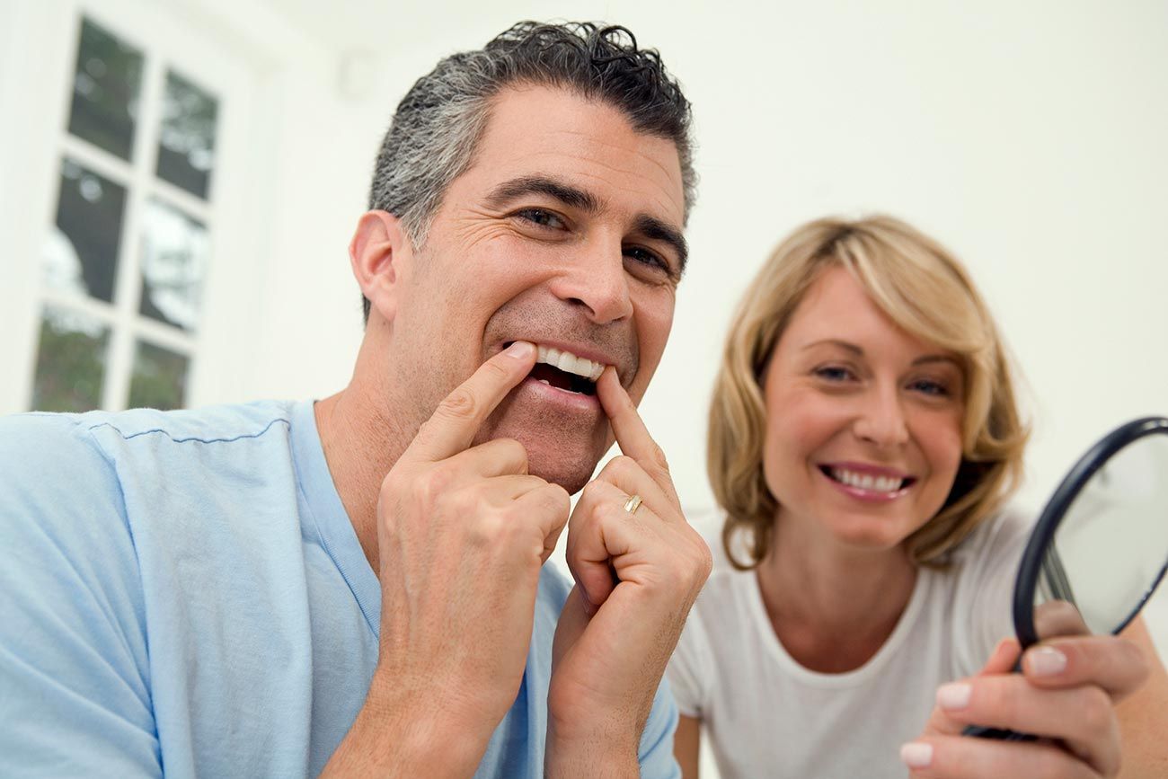 A man and a woman are looking at their teeth in a mirror.
