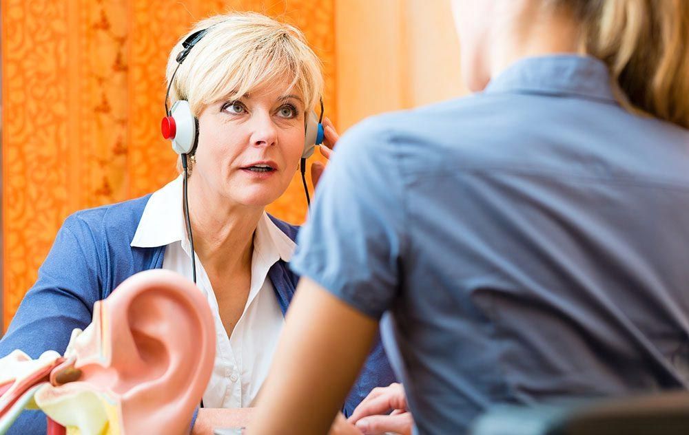 A woman wearing headphones is talking to another woman while holding a model of an ear.