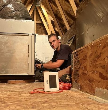 A man is sitting on the floor in an attic working on a thermostat.
