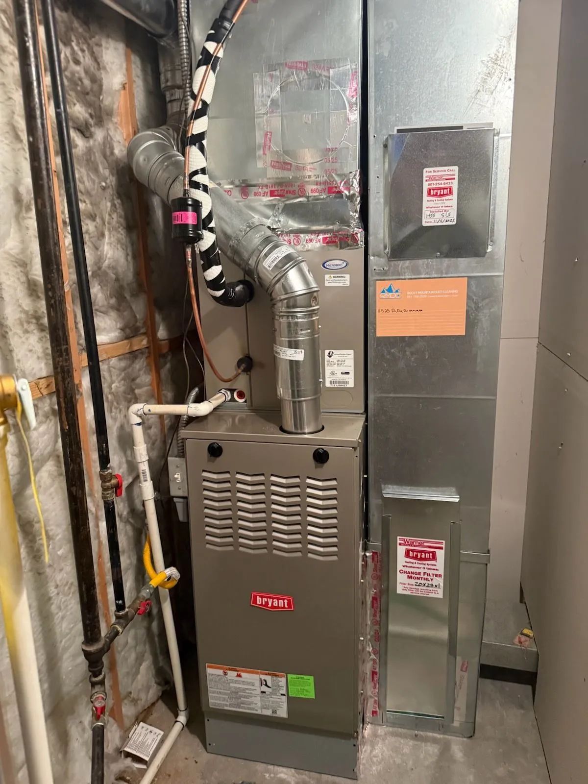Residential furnace and air handler in a utility closet with exposed pipes and ductwork.