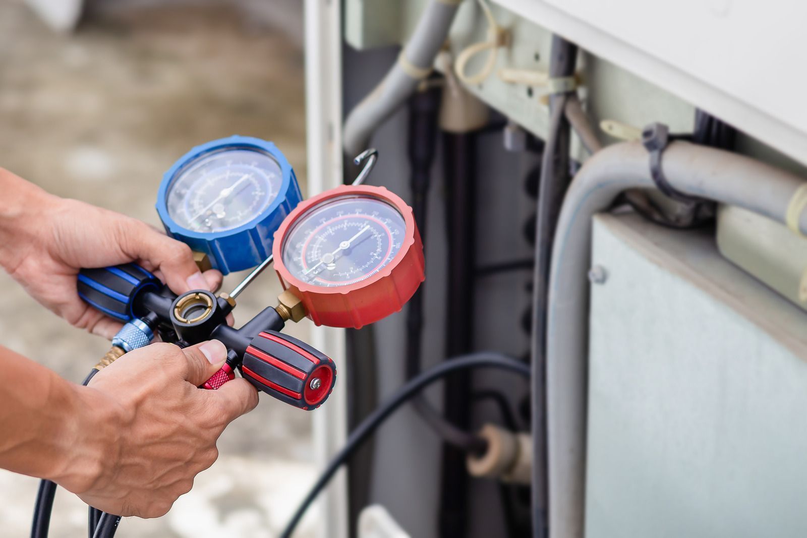 Hands holding a pressure gauge set, connected to an air conditioning unit.