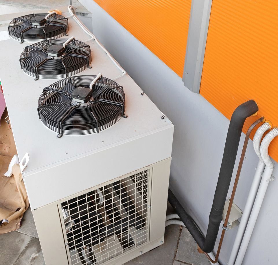 Outdoor HVAC unit with three fan blades and insulated pipes next to an orange wall.