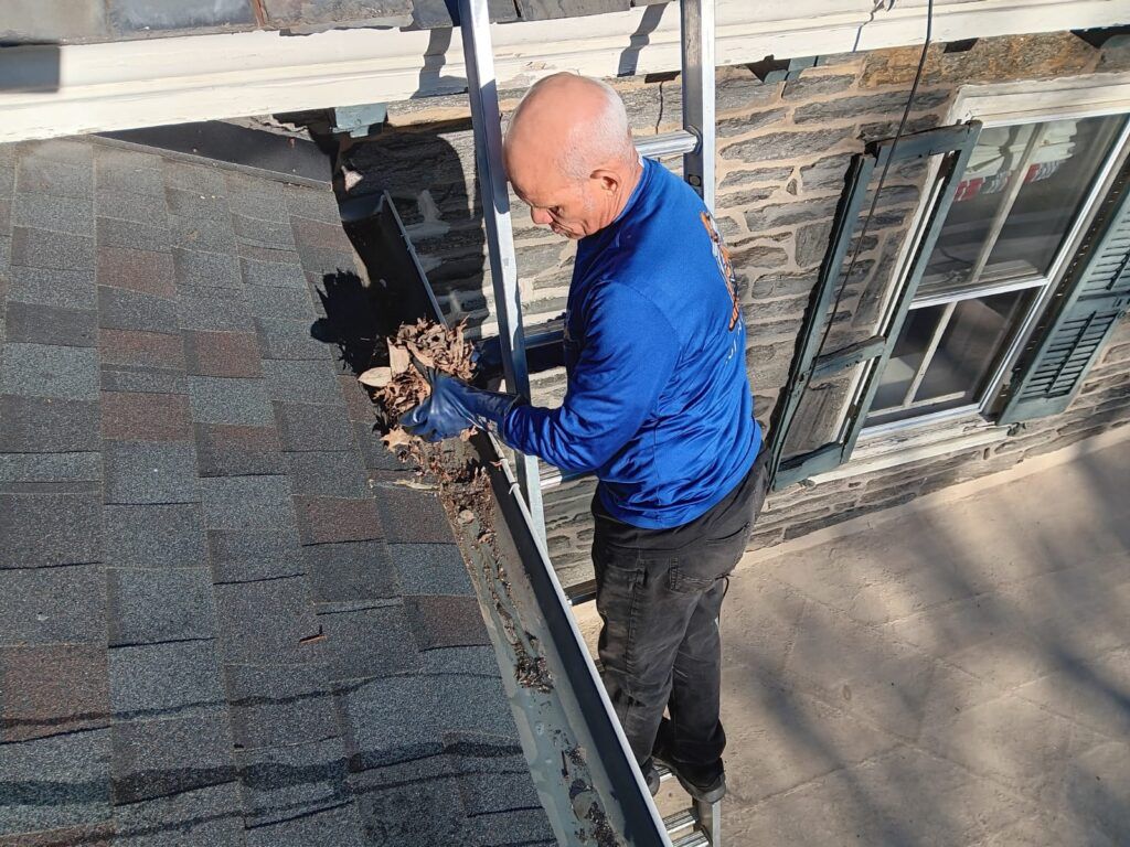 Man cleaning roof gutter with leaves, standing on a ladder. House in background.