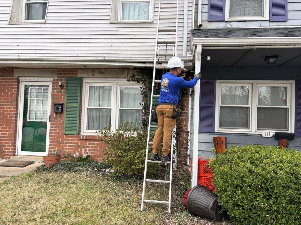 A person on a ladder working on a house gutter. Brick home with green door on the left and purple shutters on the right.