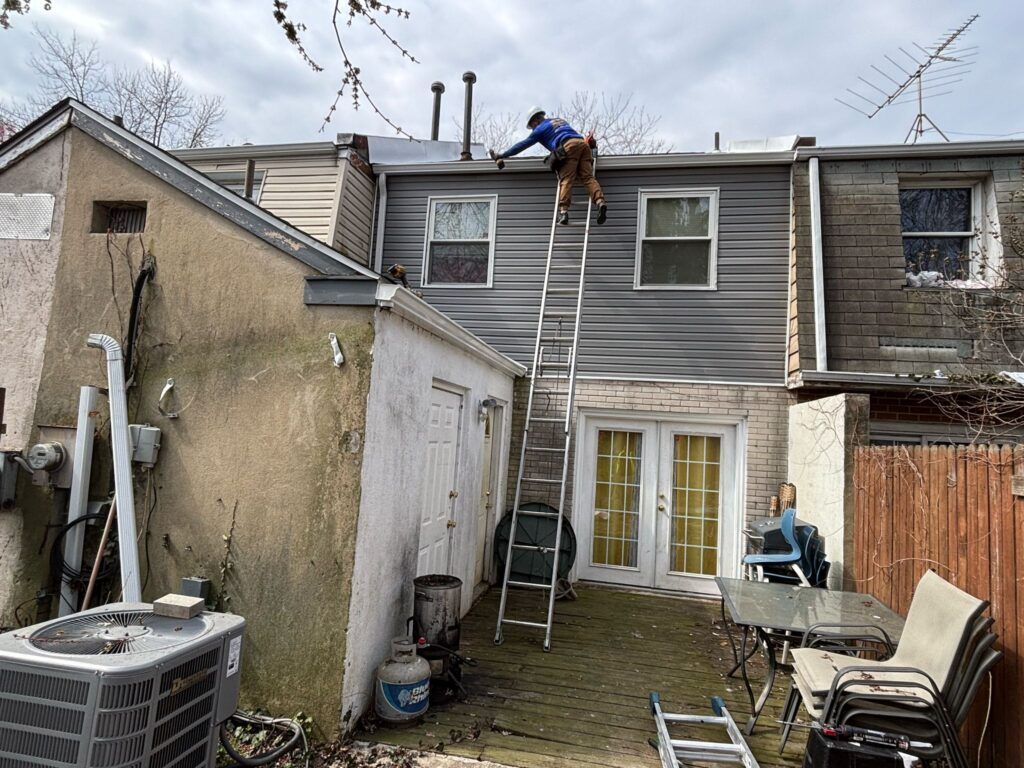 A person on a ladder working on a gray-sided building's roof; backyard setting with air conditioning unit.