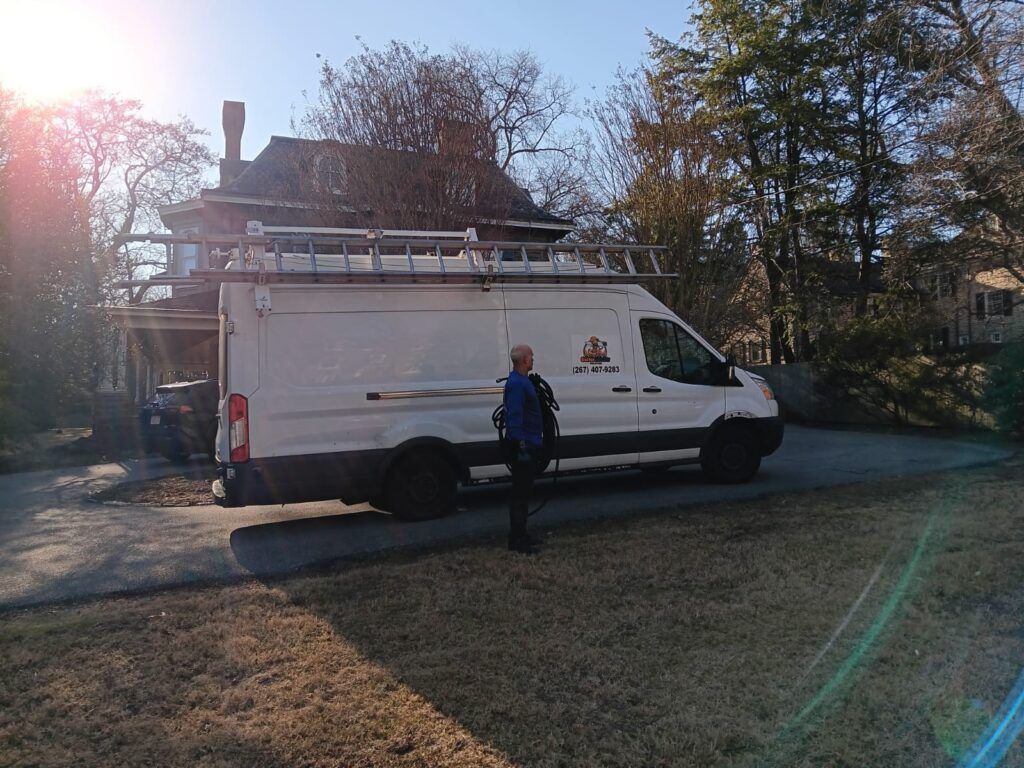 White work van parked in front of a house, with a person standing beside it. Ladder on top.