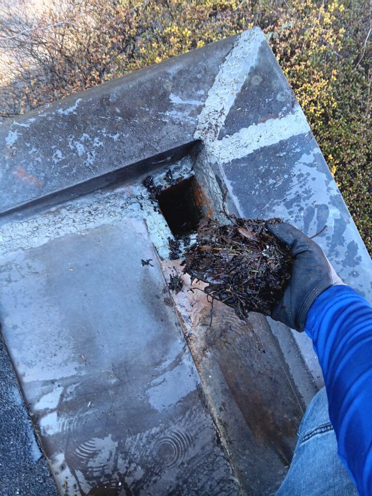Person in blue shirt and gloves cleaning a debris-filled chimney opening on a flat roof.
