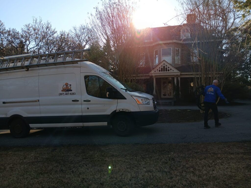 White van parked on a street near a house. A person stands nearby, gesturing. Sunlight shines.