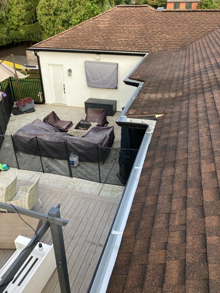 Brown-shingled roof with gutters. View of a deck, small white building, and patio with covered furniture.