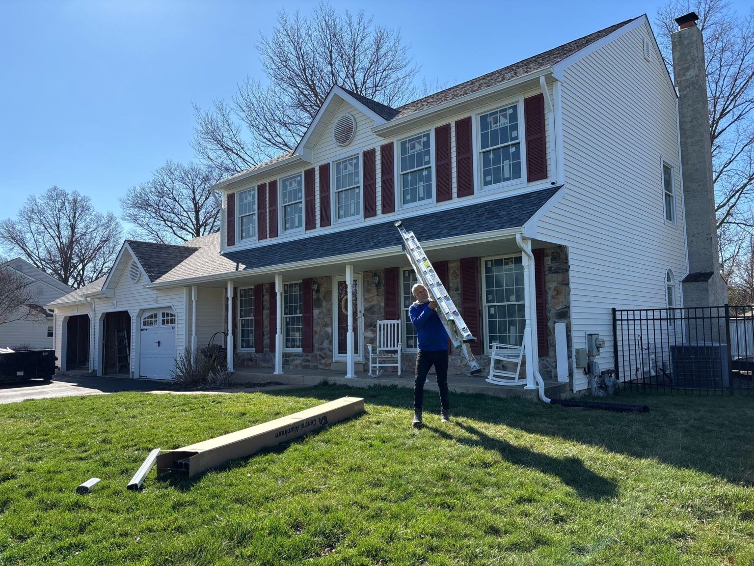 Person holding a ladder near a white house with red shutters on a sunny day.