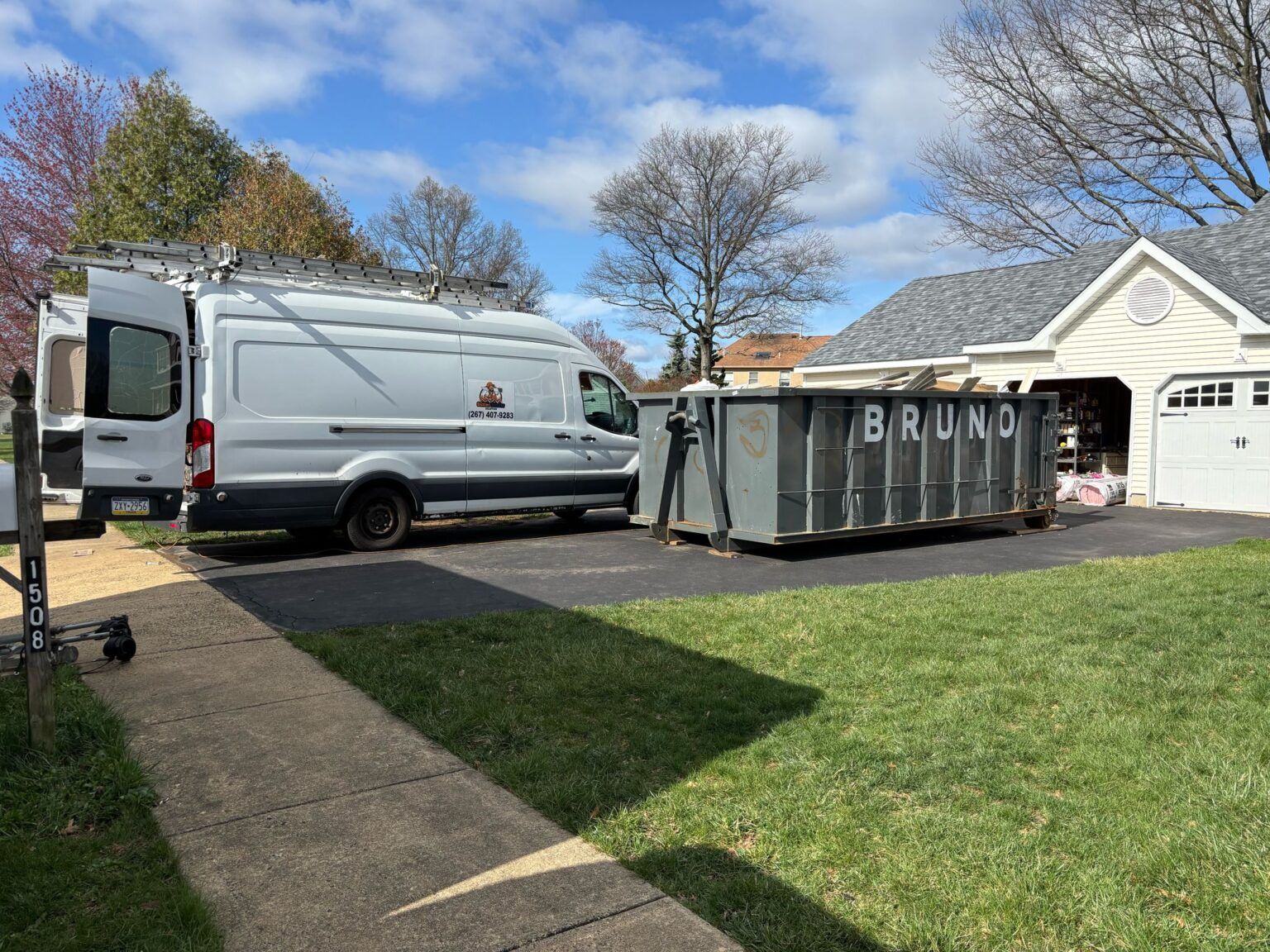 White work van parked next to a large dumpster in a residential driveway.