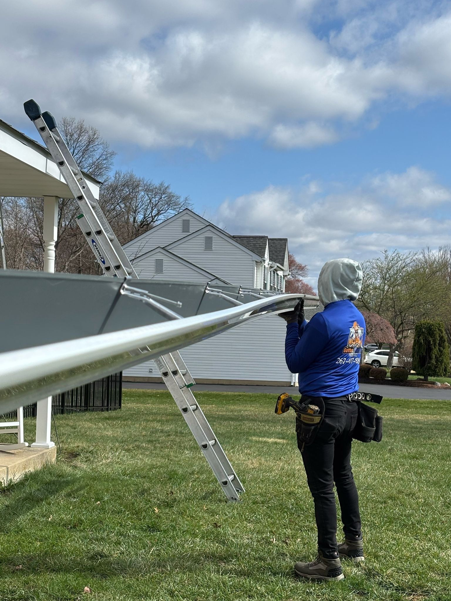 Man in blue shirt works on awning, standing on grass. Ladder leans against the house.