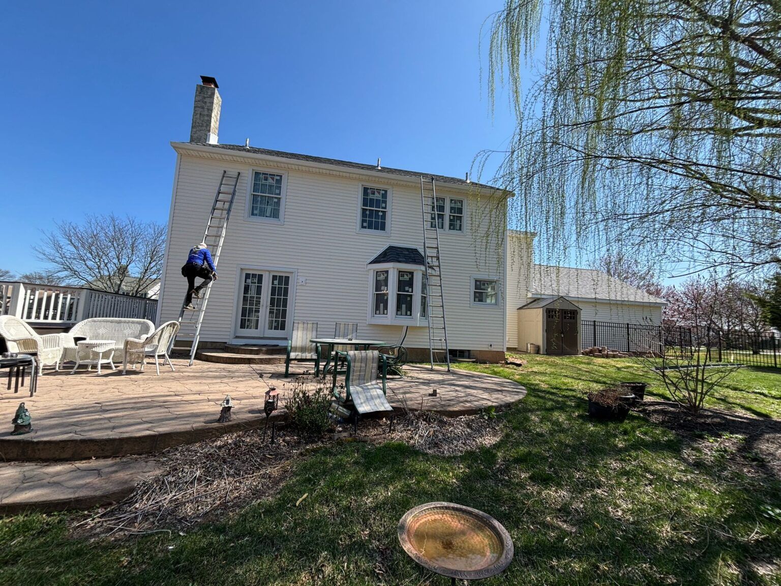 Person on ladder against a white two-story house with a chimney. Sunny day. Backyard with patio, table, and tree.
