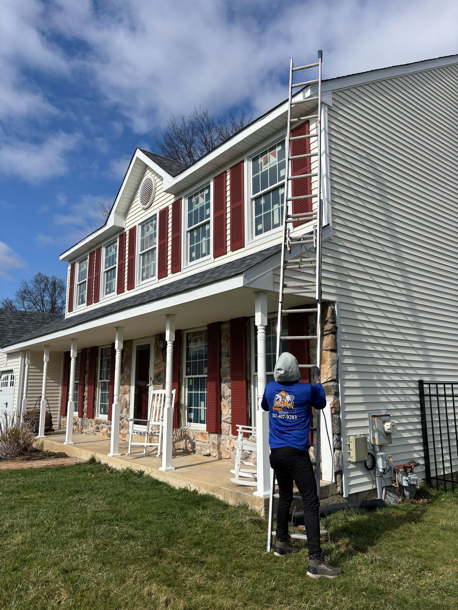 A person on a ladder working on the side of a two-story house with red shutters on a sunny day.