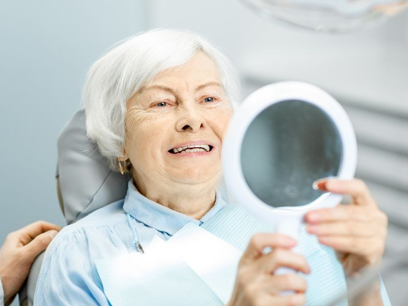 Senior woman in dentist chair looking at her teeth in a hand mirror.