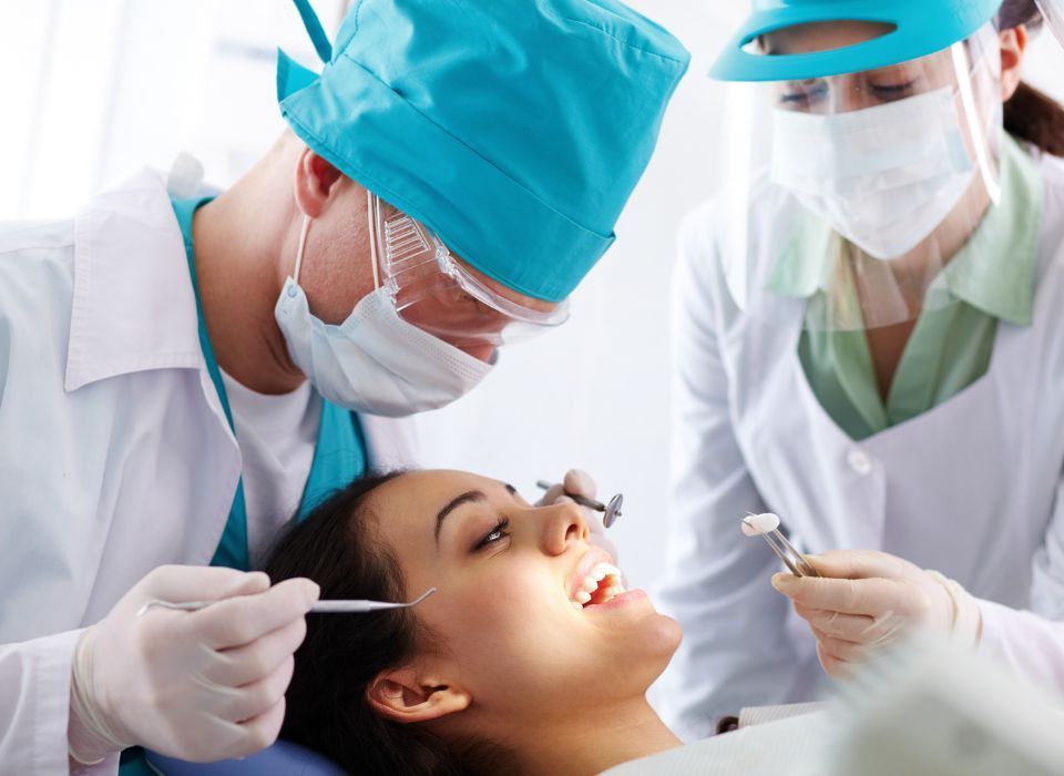 Dentist and assistant examine a patient's teeth. Bright light, white coats, dental tools.