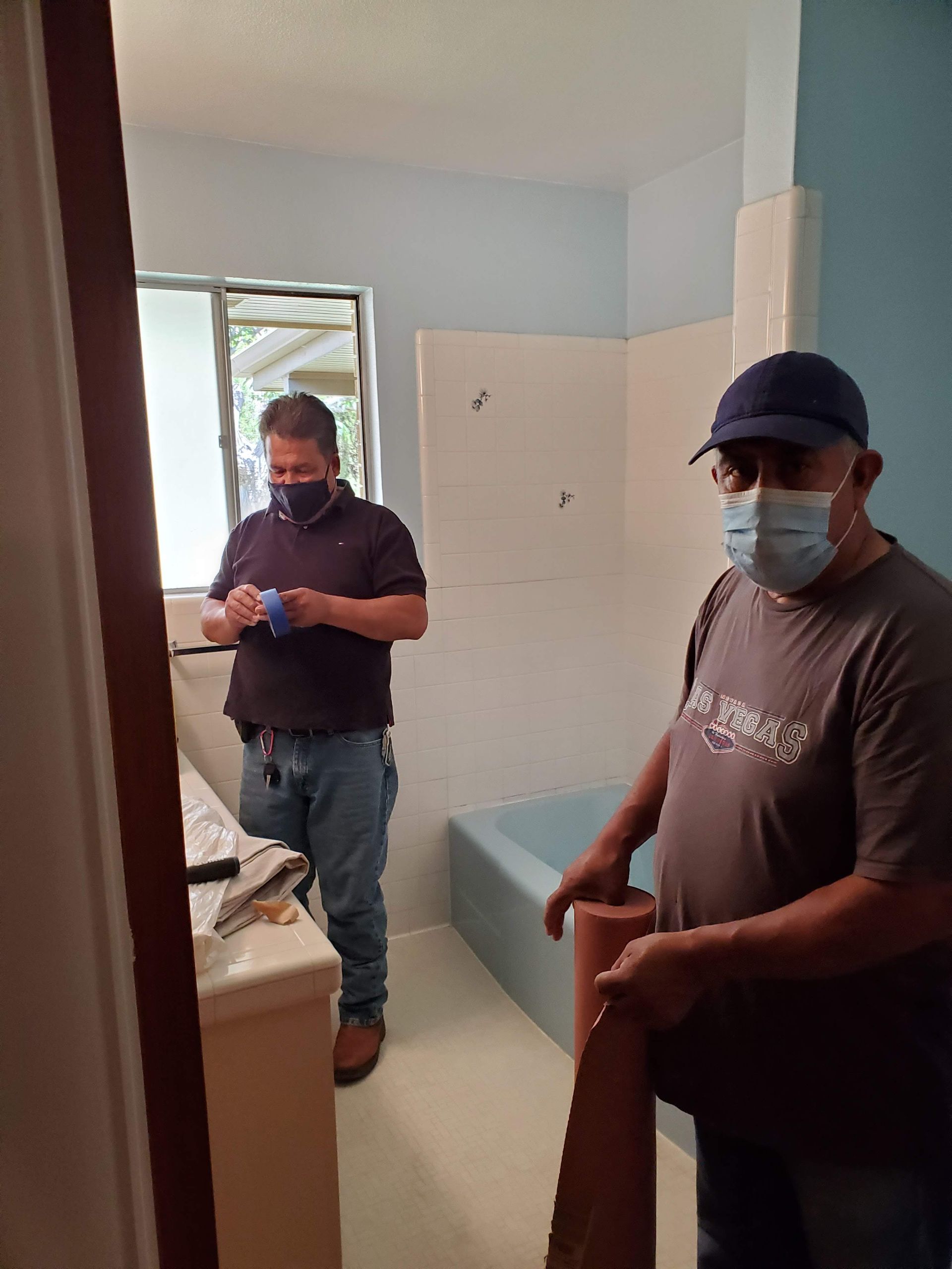 Two men wearing face masks are standing in a bathroom.