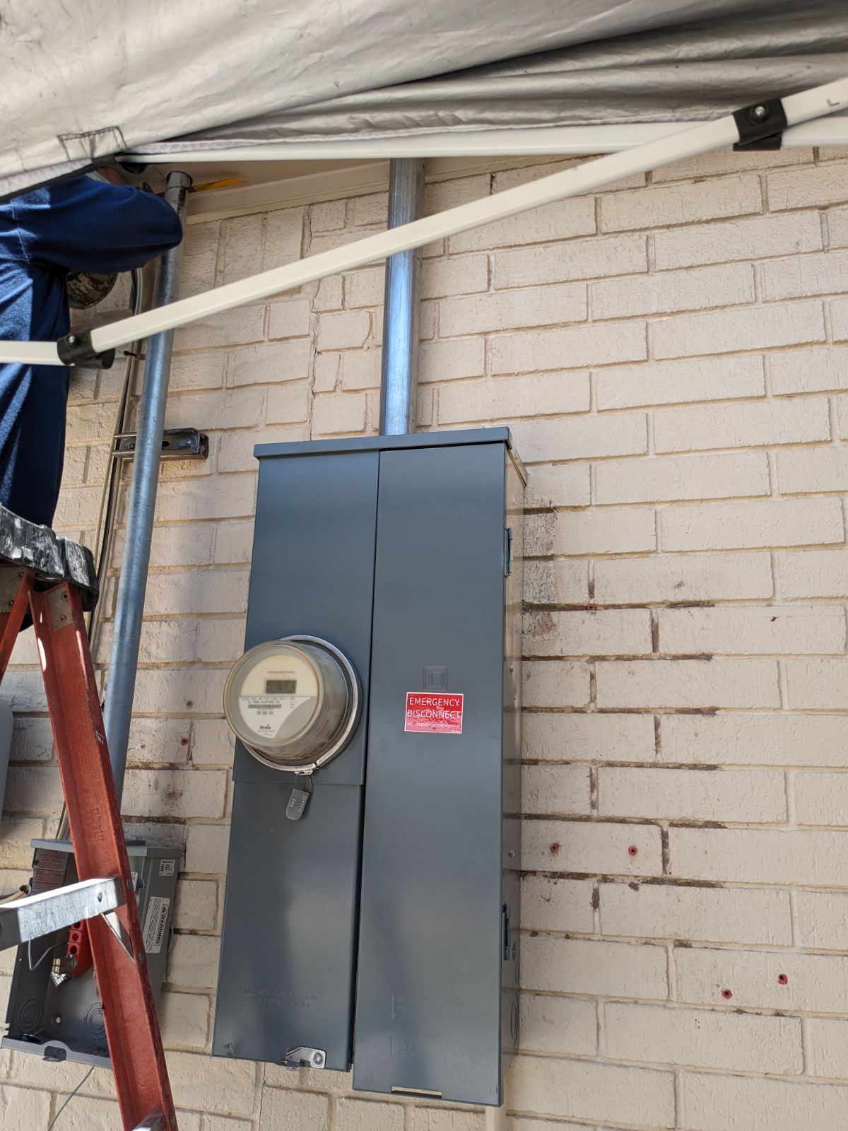 A man is standing on a ladder next to an electrical box on a brick wall.