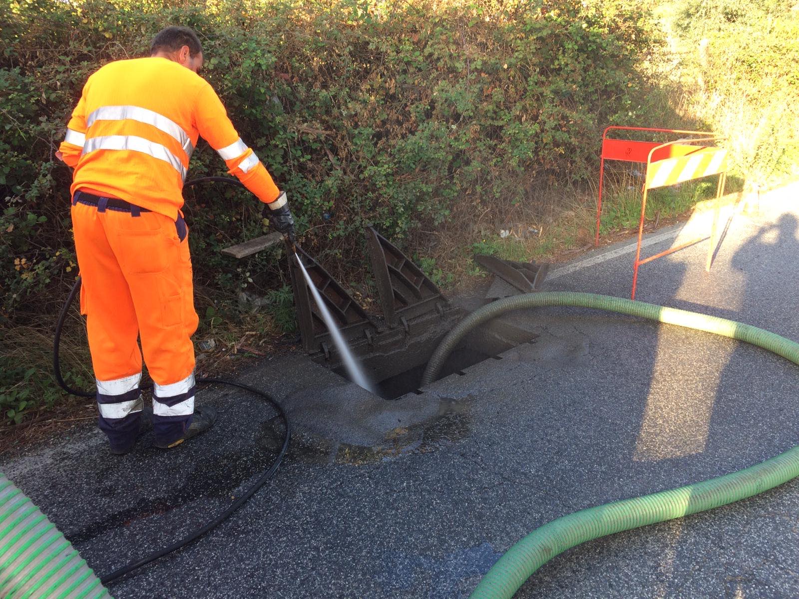 Una persona in abiti da lavoro arancioni pulisce uno scarico sul ciglio della strada con un tubo dell'acqua.