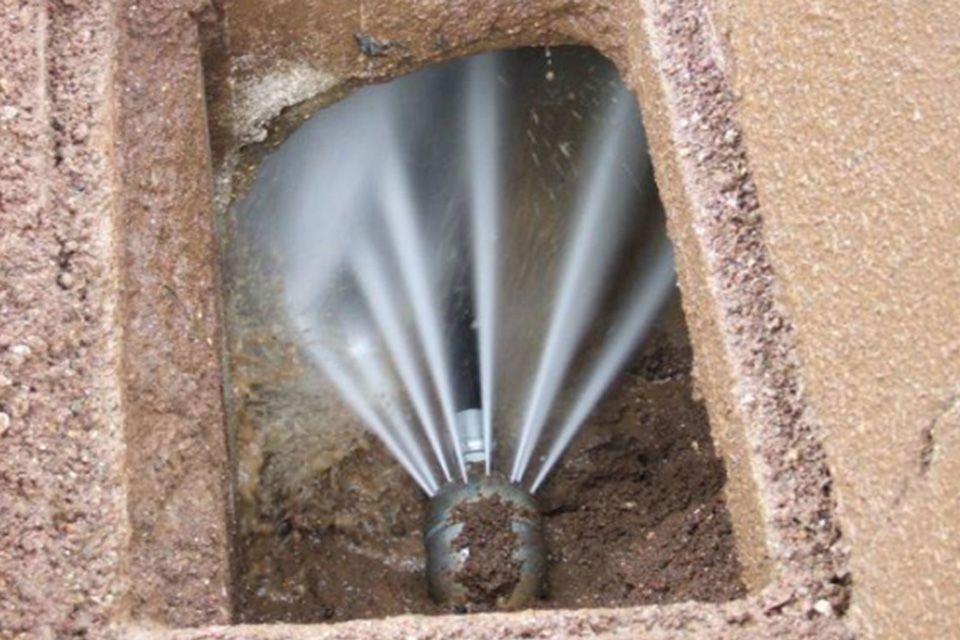 Acqua che fuoriesce con forza da un tubo sotterraneo danneggiato in un buco quadrato pieno di terra.