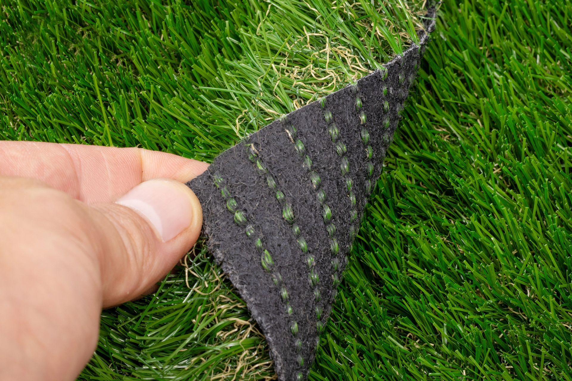 A person's hand lifts a corner of synthetic turf, revealing the black, tufted backing.