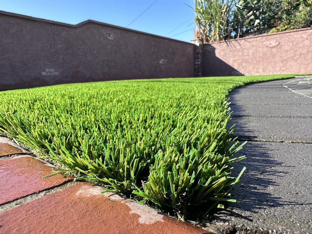 Green artificial turf lawn next to brick and gray paver paths with a brown wall in the background.