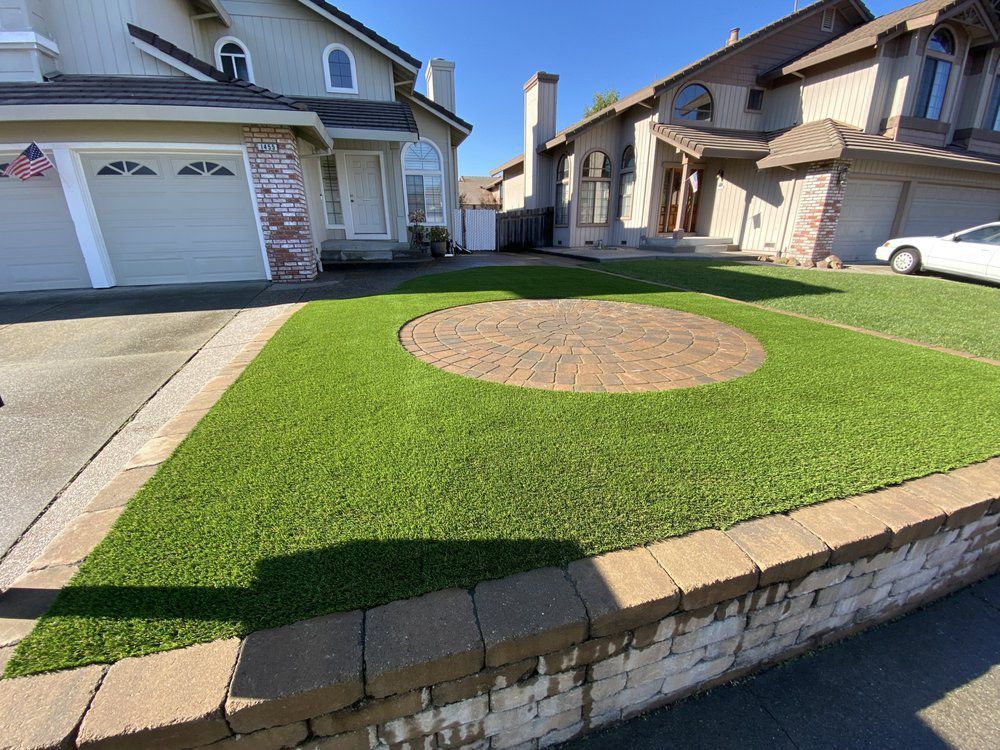 Front yard with artificial green turf, a circular paver patio, and a stone retaining wall in front of two suburban homes.