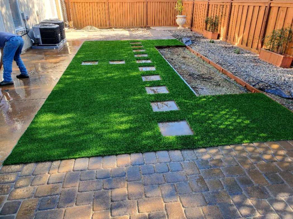 Backyard with artificial turf, stepping stones, and person working. Brown brick patio and fence.