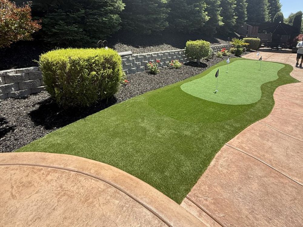A backyard putting green with artificial grass, bordered by dark gravel, a tiered stone wall, and stamped concrete paths.