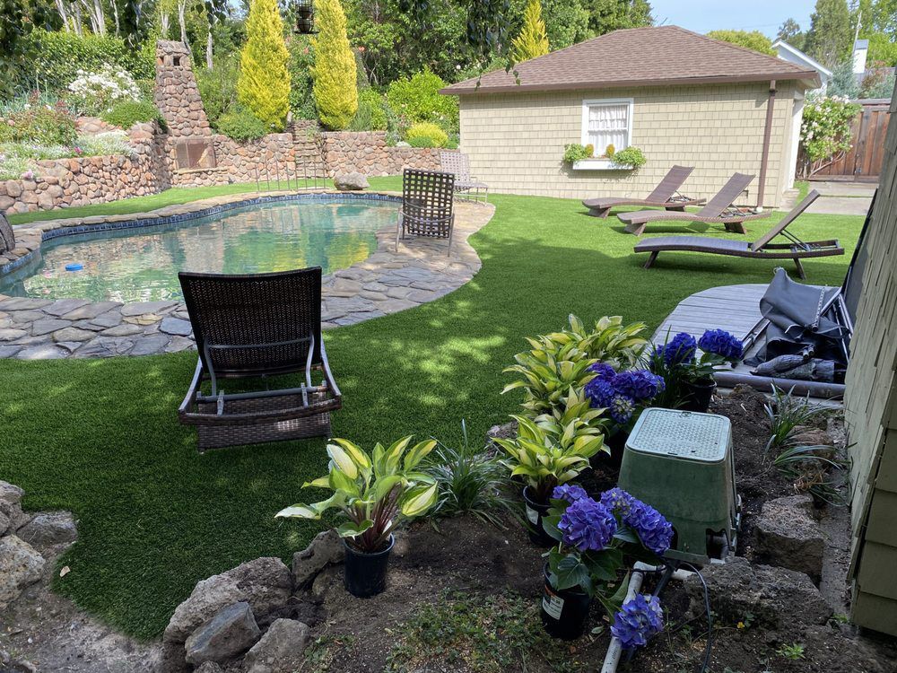A backyard with a swimming pool, stone patio, artificial grass lawn, lounge chairs, and potted flowers and shrubs.