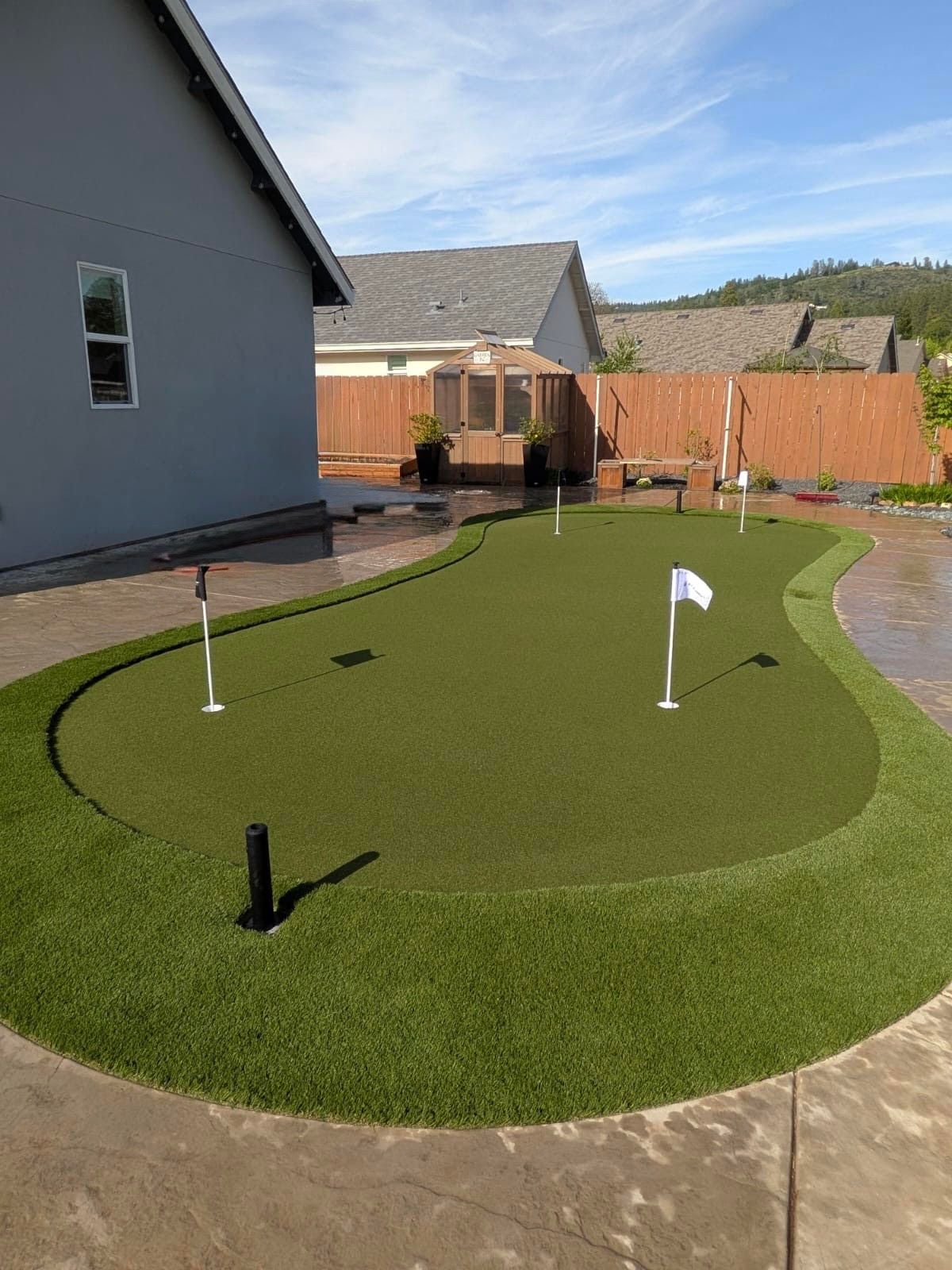 A backyard putting green with several flags, installed on a concrete patio next to a house with a wooden fence background.