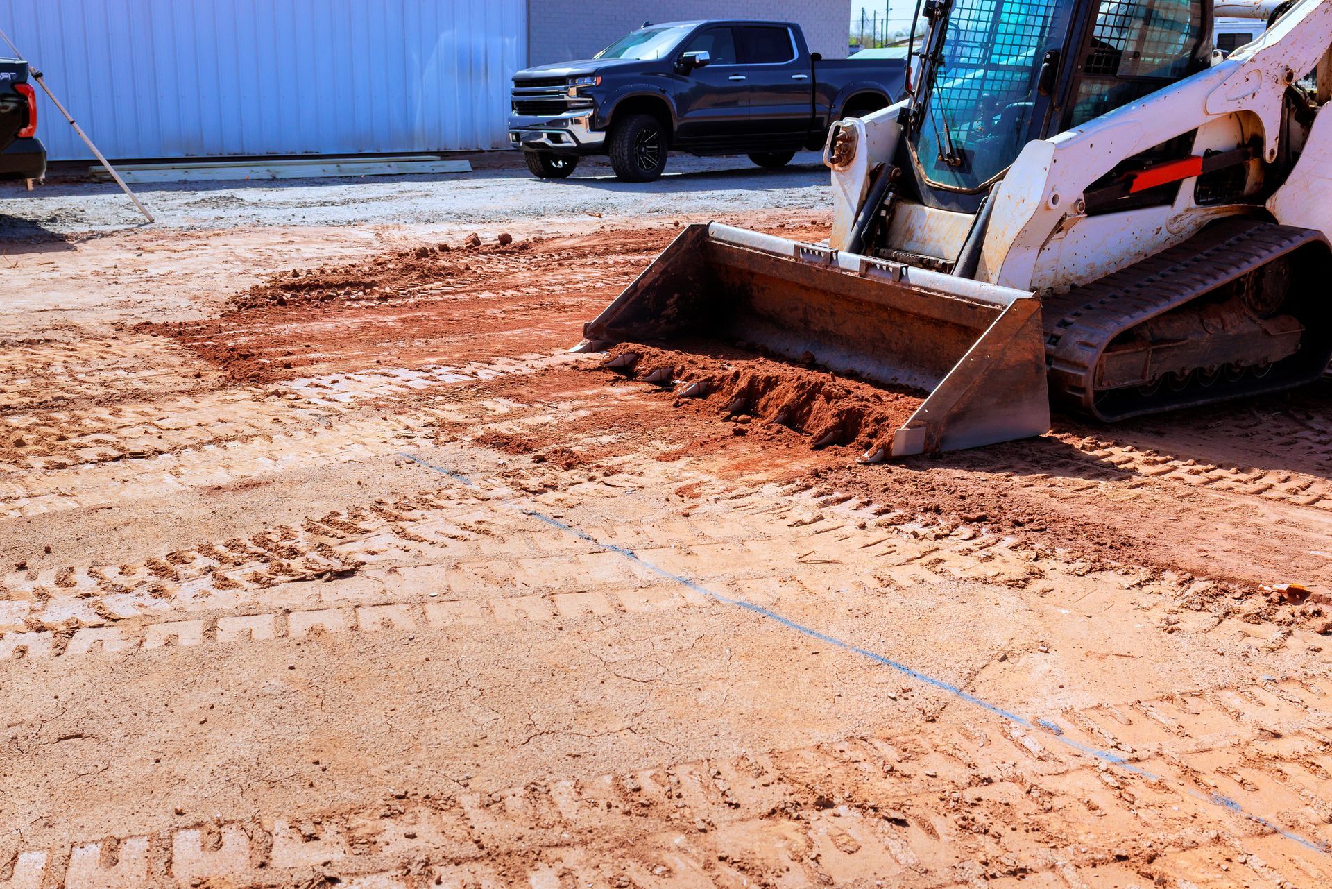 A white skid steer with a metal bucket smooths out red dirt on a construction site near a parked pickup truck.