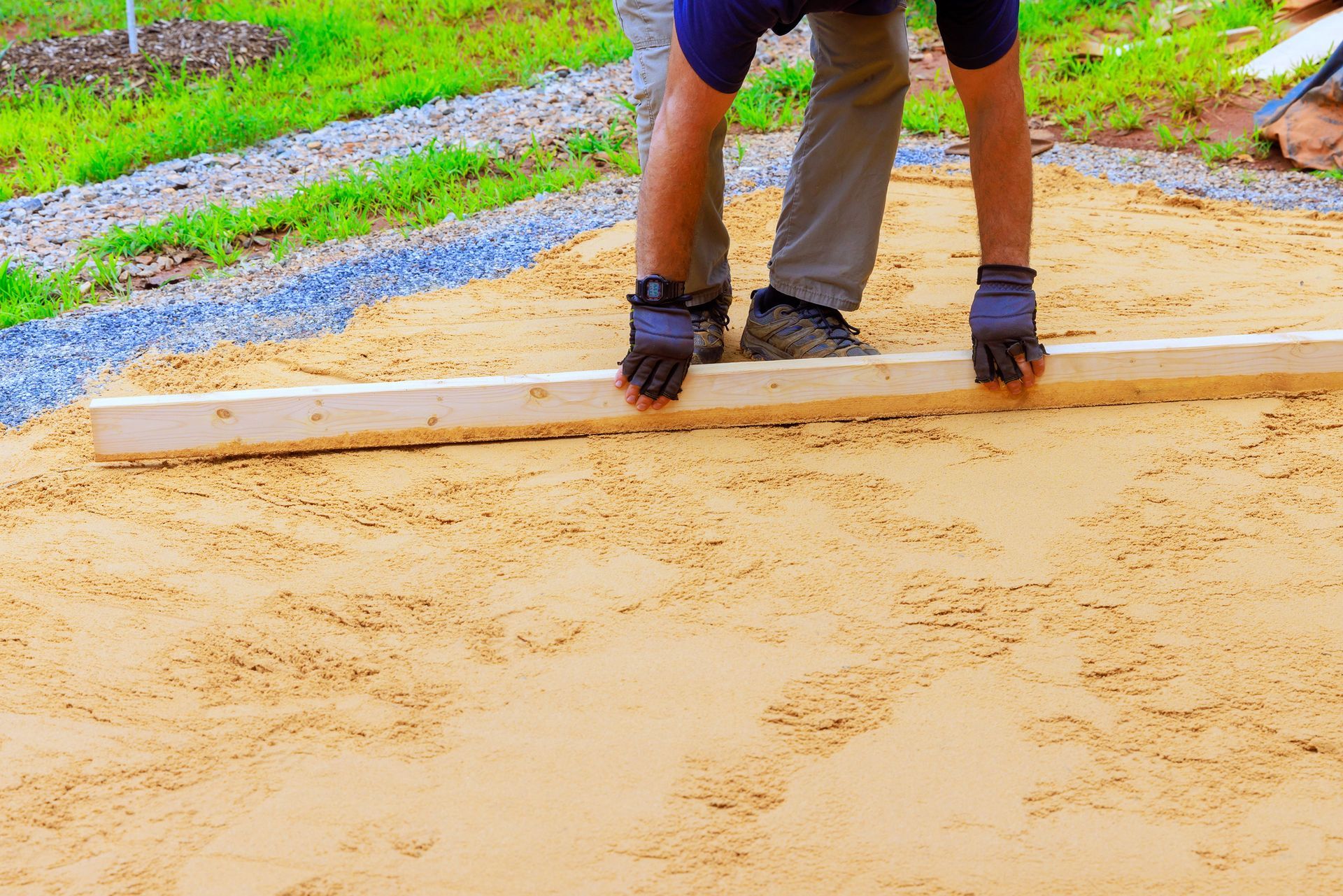 A person wearing work gloves levels sand for a patio using a long, straight wooden board.