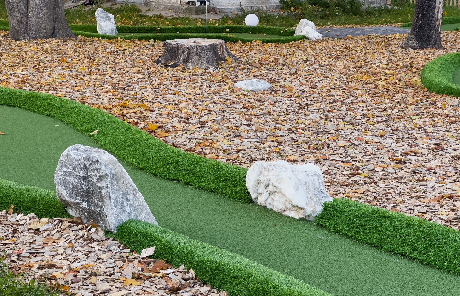 Miniature golf course green with artificial grass borders, scattered with fallen leaves, logs, and decorative rocks.