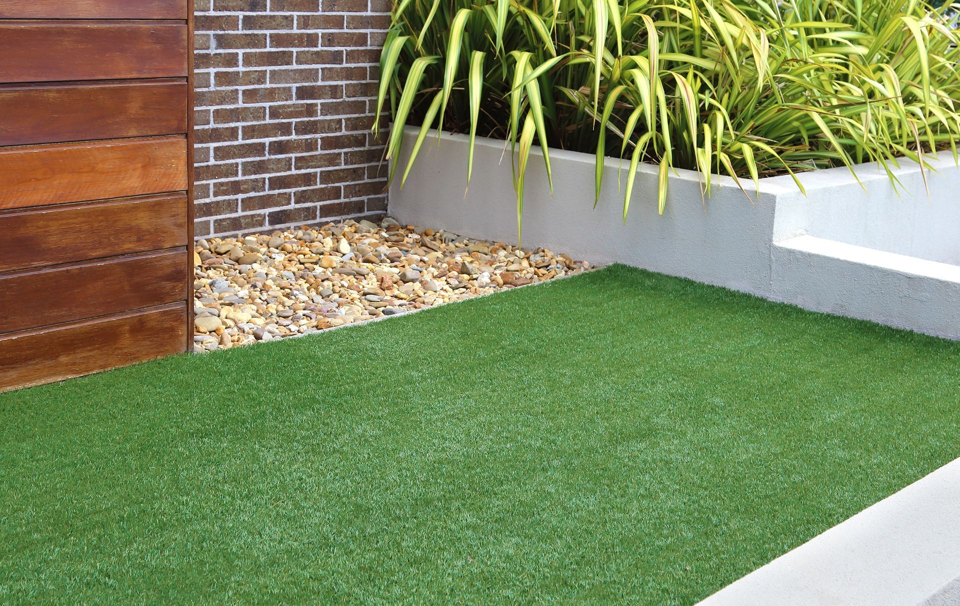 A landscaped garden corner with artificial turf, a gravel patch, a brick wall, timber fencing, and a concrete planter.