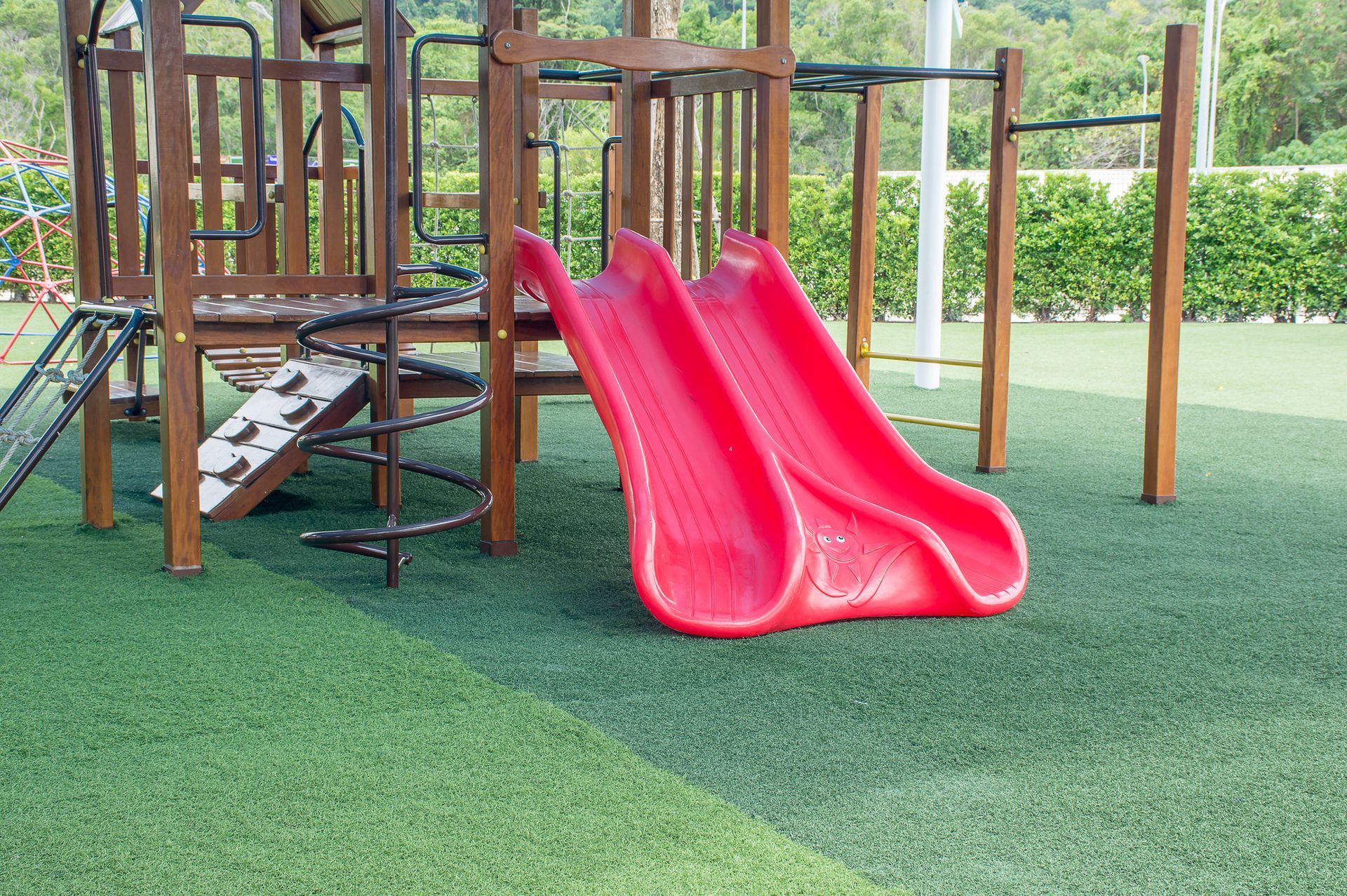 A wooden playground structure with two red plastic slides sitting on a green turf field.