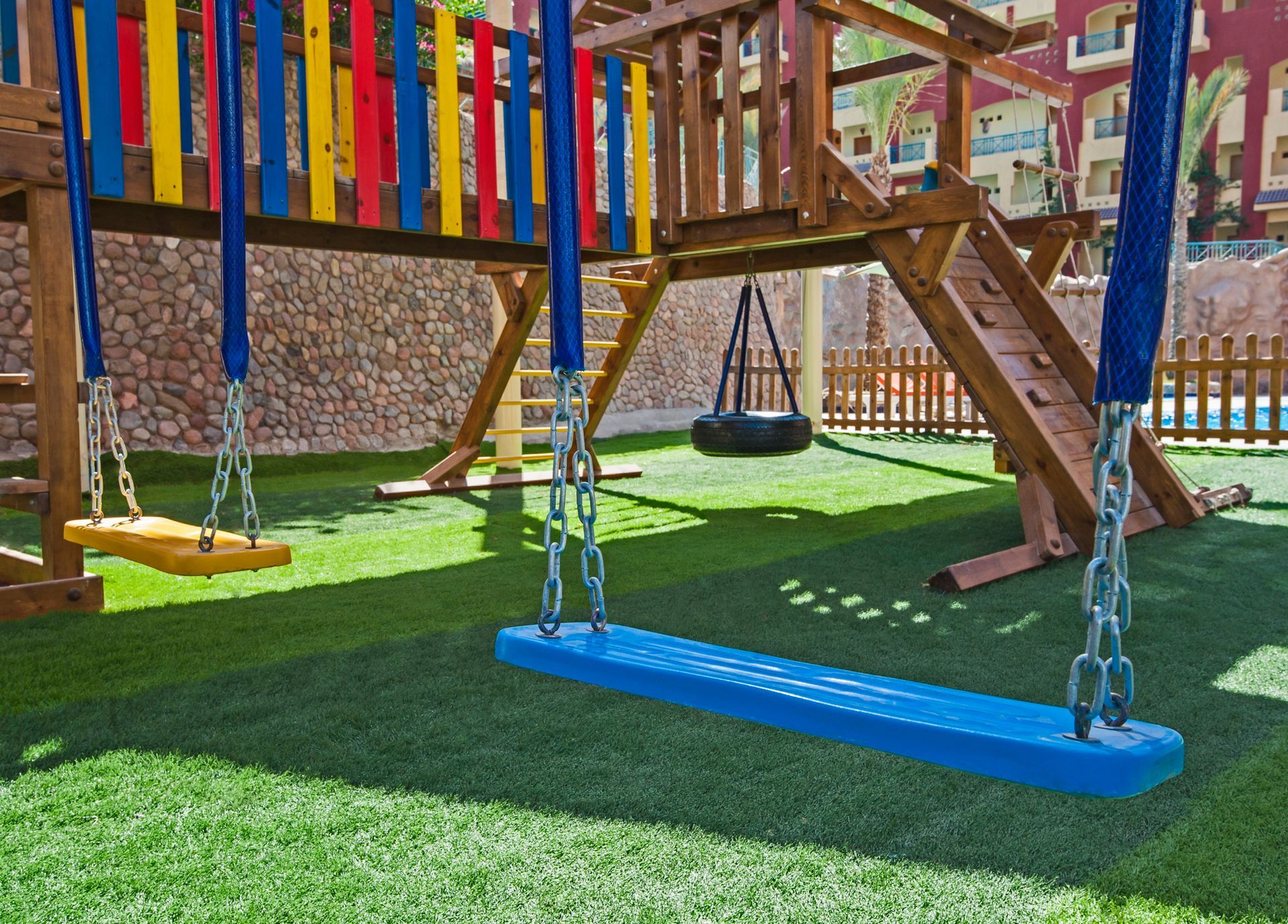 A playground with a blue swing, a yellow swing, and a tire swing hanging from a wooden climbing structure on green turf.
